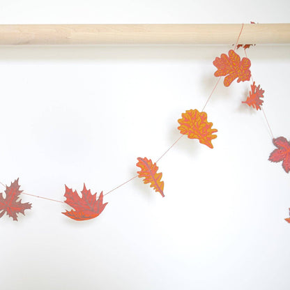 A string garland of autumn leaf cutouts in red, orange, and yellow, hanging from a light wood dowel against a plain white wall.