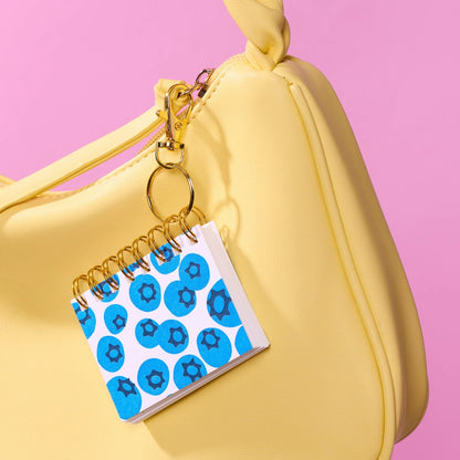 A small spiral-bound notebook charm with a blueberry patterned cover hangs from a gold clasp attached to a yellow handbag, set against a solid pink background.