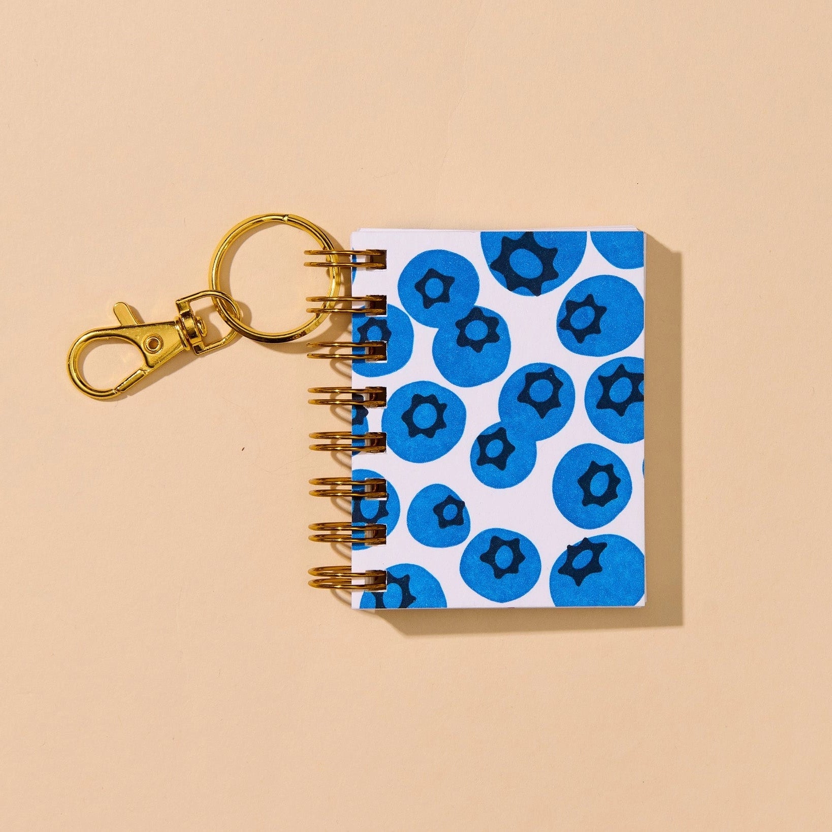 A small spiral-bound notebook keychain with a white cover decorated with blueberry motifs, attached to a gold-tone metal clasp and key ring, shown on a solid beige background.