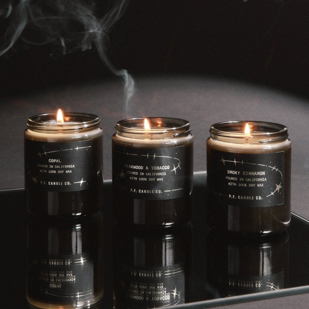 Three dark grey glass jar candles with black labels are lit and displayed on a reflective black surface, with smoke drifting above them against a dark background.