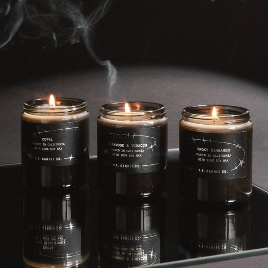 Three dark grey glass jar candles with black labels are lit and displayed on a reflective black surface, with smoke drifting above them against a dark background.