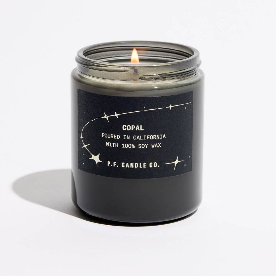 A lit scented candle in a dark gray glass jar with a black label reading 'COPAL POURED IN CALIFORNIA WITH 100% SOY WAX' and ‘P.F. Candle Co'
