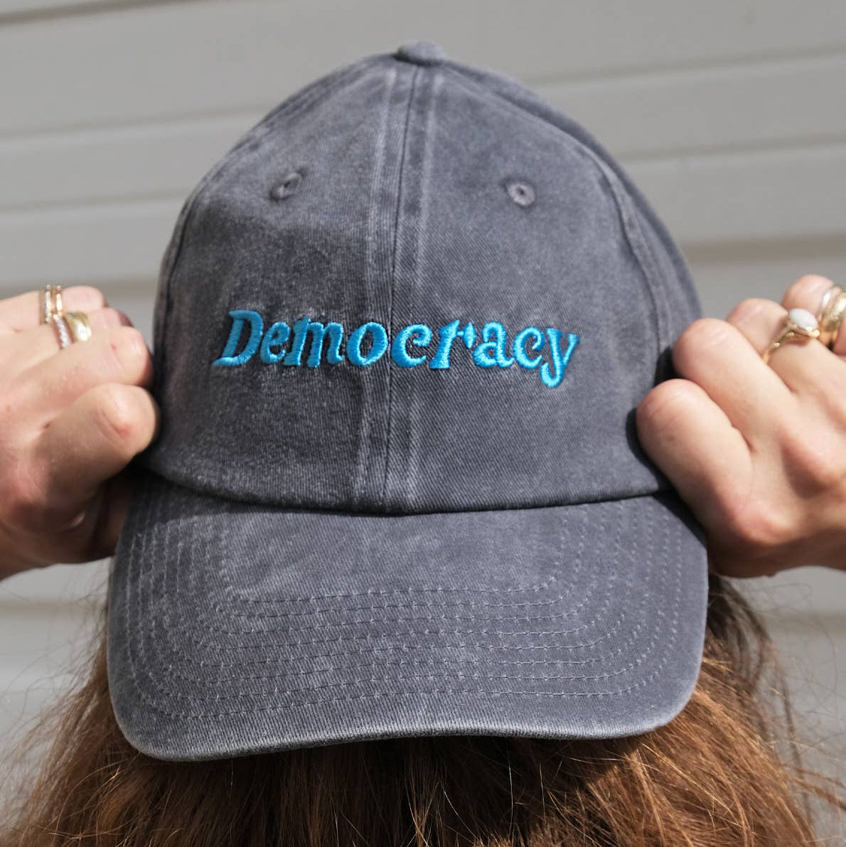 Gray cap with 'Democracy' embroidered on it held by a person against a neutral background