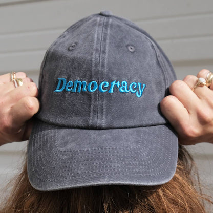 Gray cap with 'Democracy' embroidered on it held by a person against a neutral background