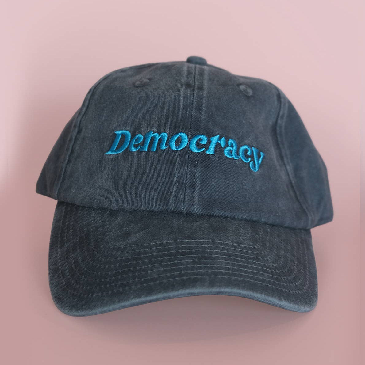 A washed dark blue baseball cap with the word 'Democracy' embroidered in bright blue script across the front, set against a solid pink background.