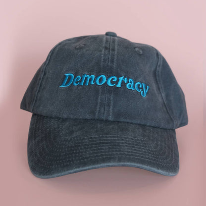 A washed dark blue baseball cap with the word 'Democracy' embroidered in bright blue script across the front, set against a solid pink background.