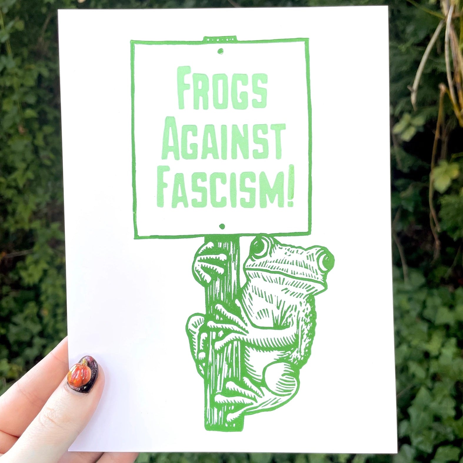 Print with 'Frogs Against Fascism!' text held against a green leafy background