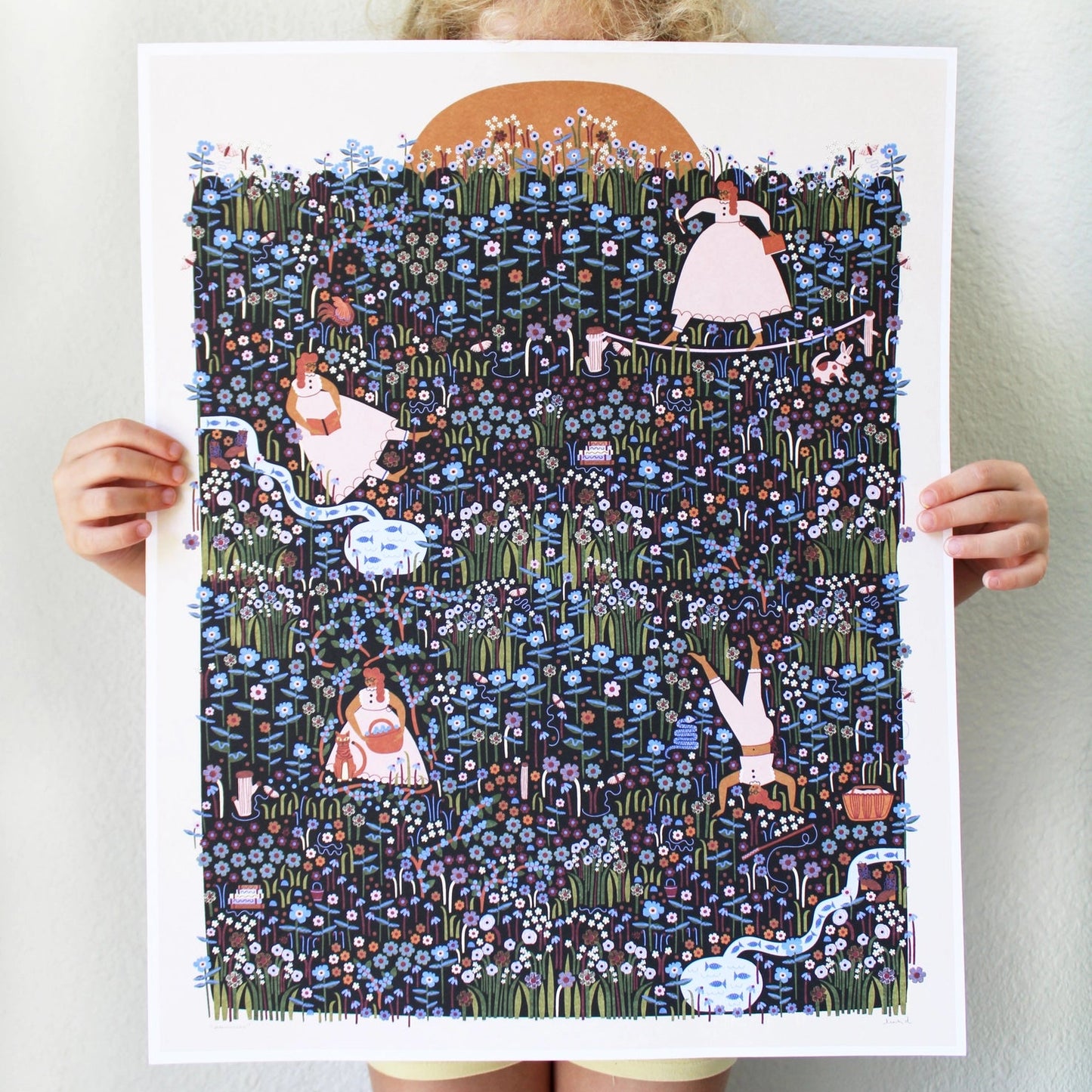 A person holding up a large white art print featuring a whimsical illustration of a dark blue wildflower field with small figures, winding light-blue streams, and a tan sun. 