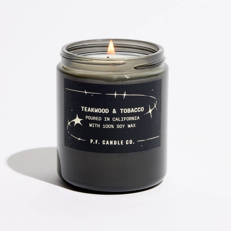 A lit candle in a dark glass jar with a black label reading 'TEAKWOOD & TOBACCO' and ‘P.F. Candle Co'