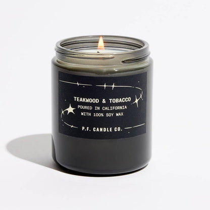 A lit candle in a dark glass jar with a black label reading 'TEAKWOOD & TOBACCO' and ‘P.F. Candle Co'