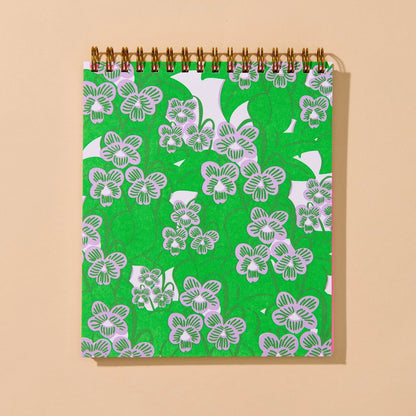 A spiral-bound notebook with a bright green cover featuring a white floral pattern, shown against a solid beige background.
