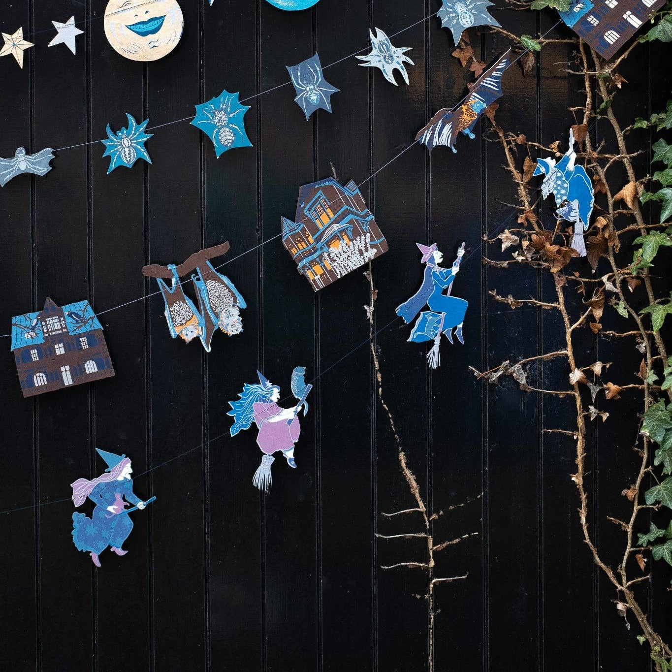 A decorative garland made of blue and white paper cutouts, including stars, moon phases, houses, and whimsical characters, hung against a dark wooden wall with ivy on the right side.