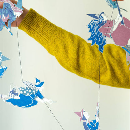 A person wearing a chunky mustard yellow knit sweater holds up a string of colorful paper witch cutout figures against a plain light background.