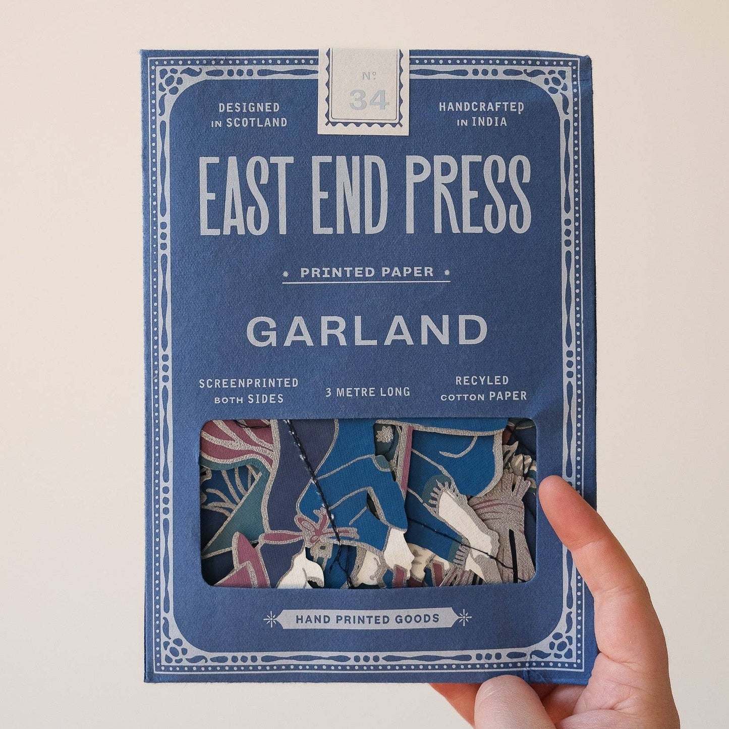 A hand holding a blue package of East End Press Garland printed paper against a plain light background. The package features white text and a window showing the colorful patterned sheets inside.