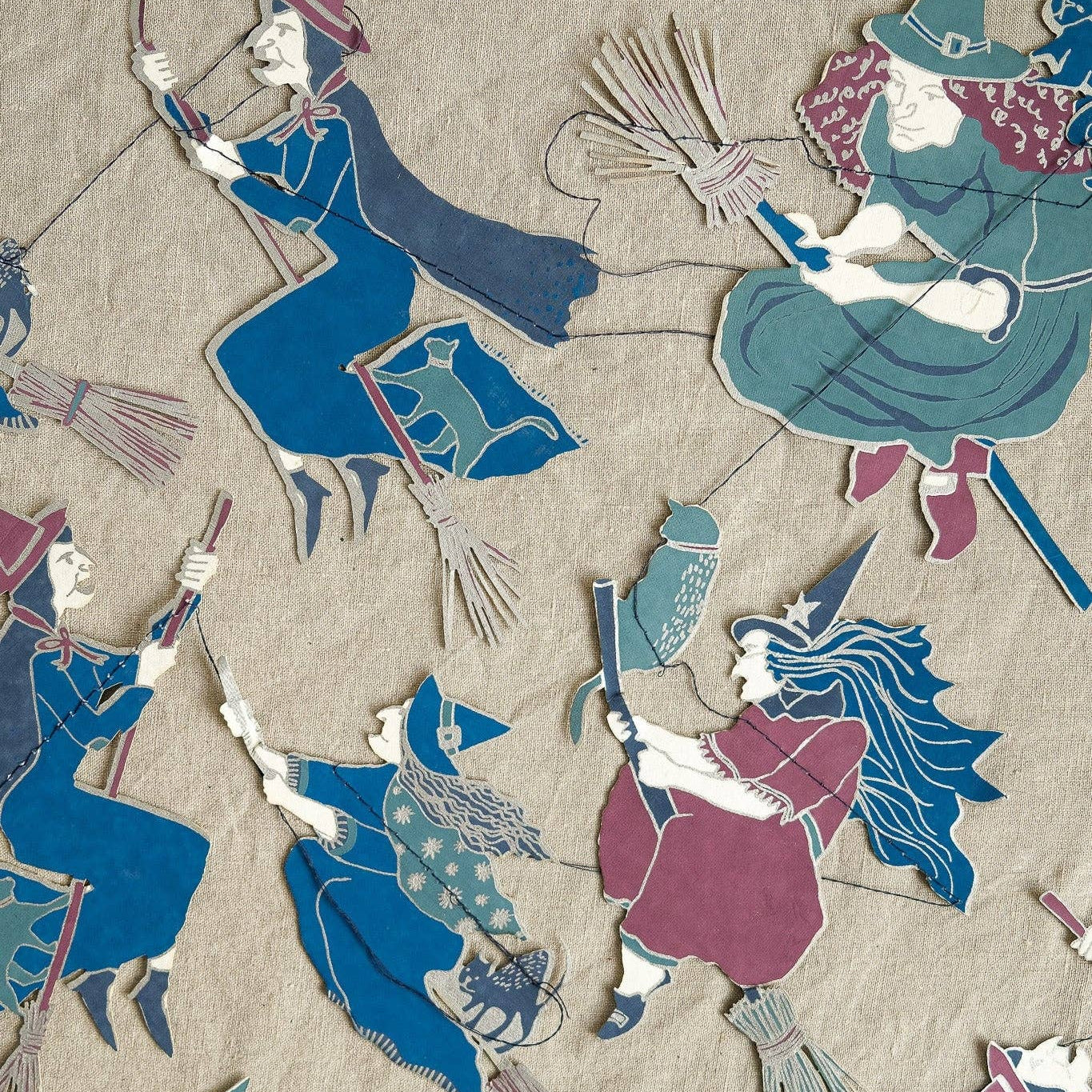 A close-up of a garland laid flat featuring multiple stylized witch figures in blue, teal, burgundy, and cream on a beige background.